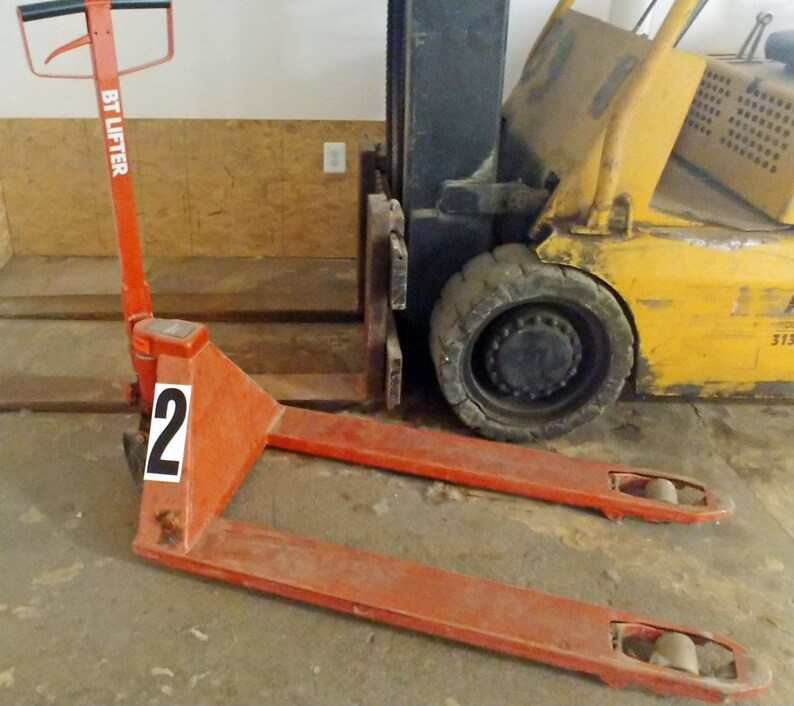 A red manual pallet jack labeled BT Lifter with a number 2 sticker is positioned on the floor next to a yellow forklift inside a warehouse.