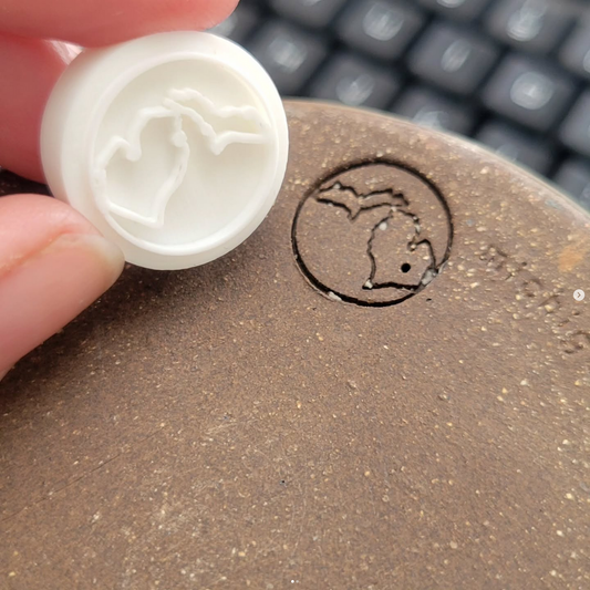 Hand holds a Pottery Makers Mark Stamp, 3 round, for pottery; features Michigan Upper and Lower Peninsula design for Michigan Clay. Stamp is being held next to a fired mug showing the depth and clarity of the stamp and design