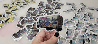 Die cut vinyl sticker of a graffiti truck by InkBird Print Studio, held by hand with black nails; custom stickers on white background.