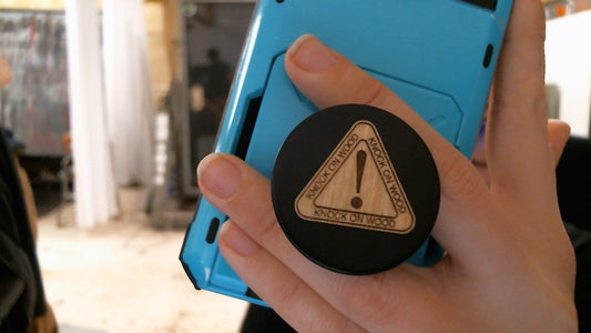 Hand holding blue phone with InkBird Print Studio vinyl Knock On Wood pop socket—black, triangle, exclamation mark, bold text.