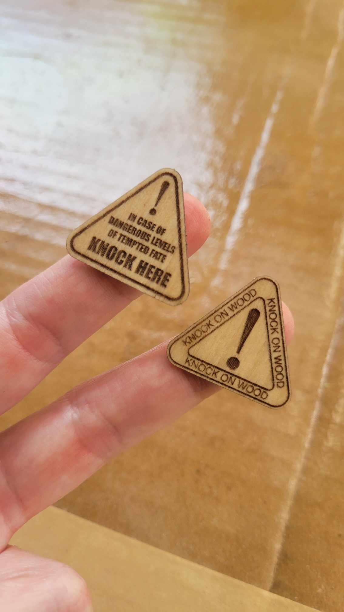 A hand holds two small, triangular wood veneer stickers by InkBird Print Studio engraved with phrases and exclamation marks. "Knock on Wood" stickers